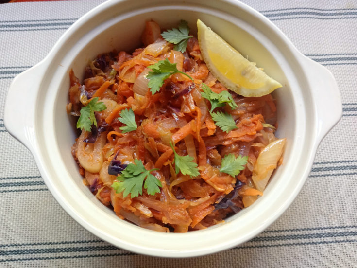 Braised cabbage with tomato in a cream-coloured casserole dish, garnished with fresh parsley and a lemon wedge on a striped cloth background.