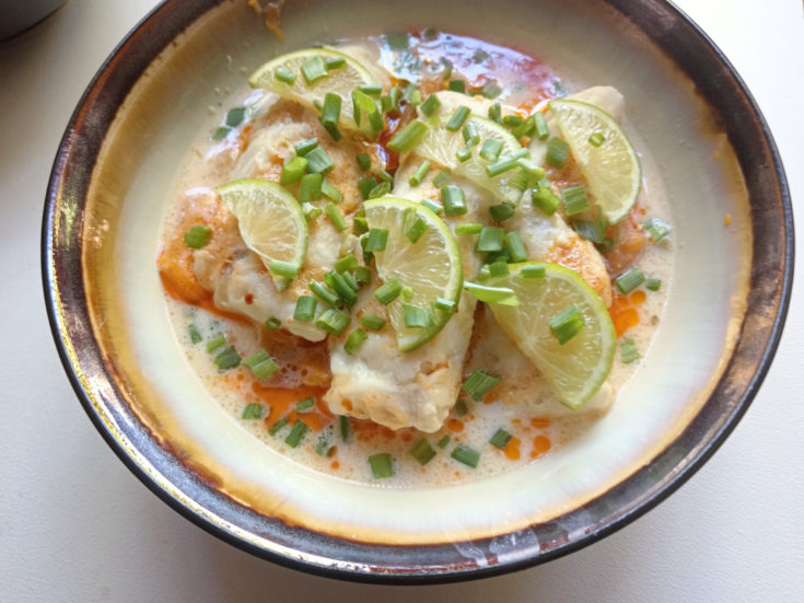 Poached hake medallions in coconut-lime broth, topped with smoked paprika butter, lime slices, and spring onions in a ceramic bowl.